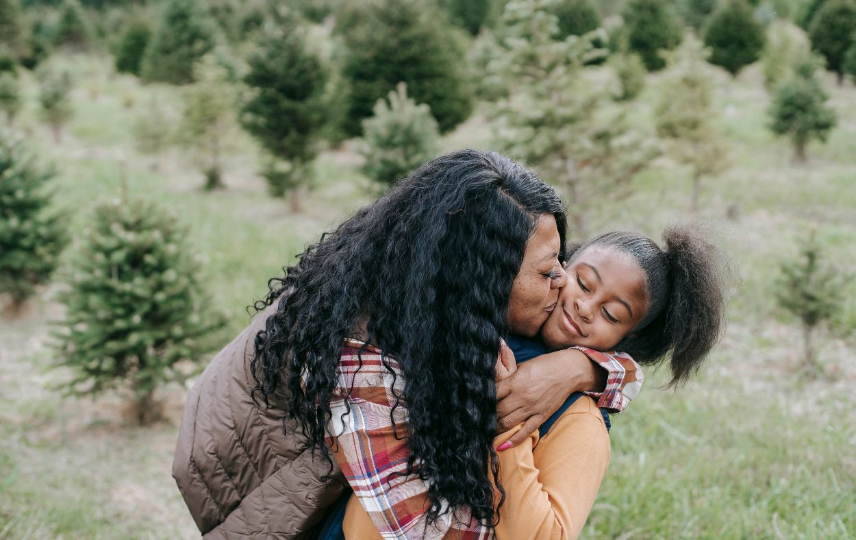 Mom hugging and kissing her daughter outside with trees in the background